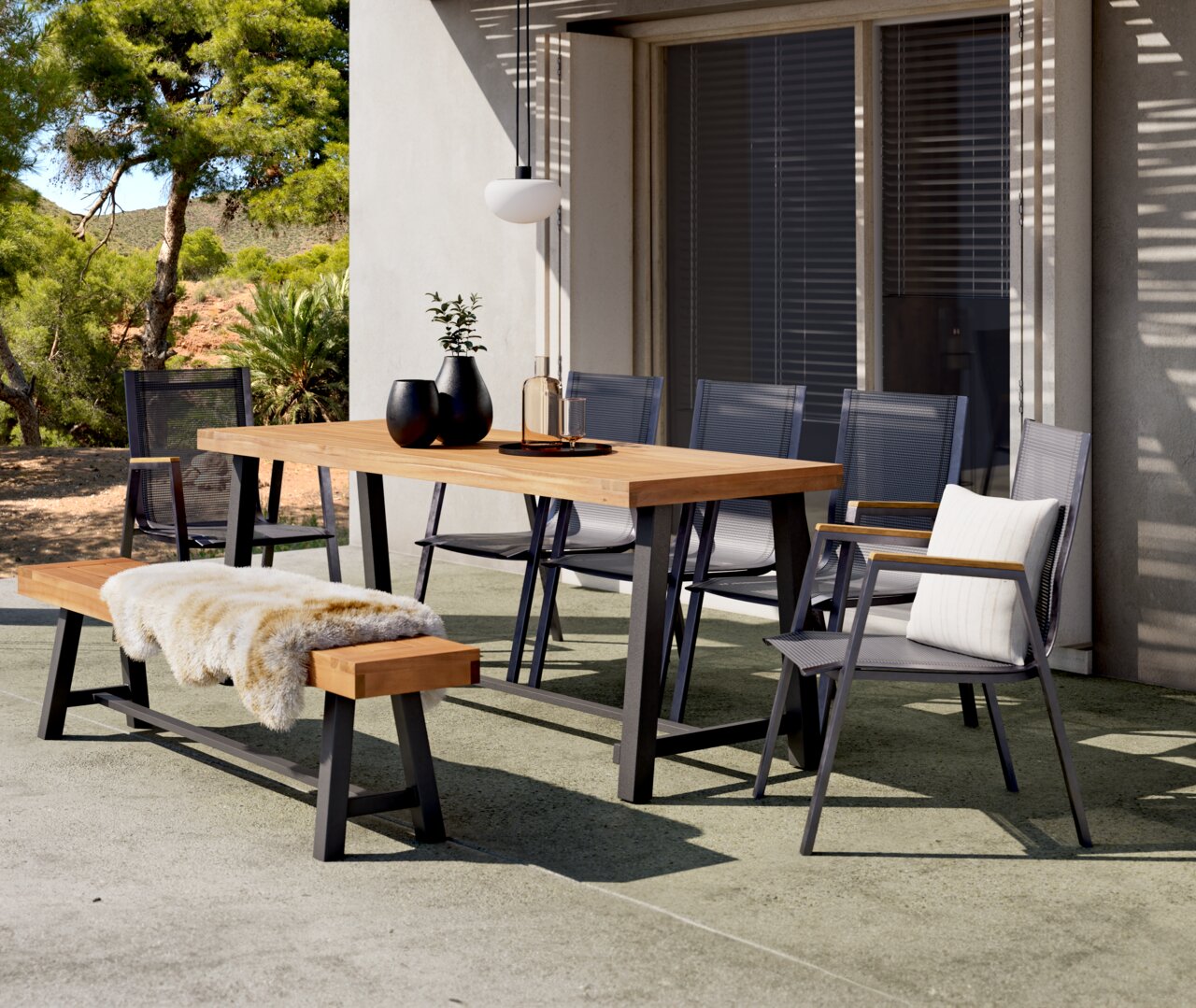 Moderner Gartentisch aus Holz mit Metallgestell, umgeben von Stühlen und einer Bank, auf einer Terrasse mit Blick auf einen Garten.