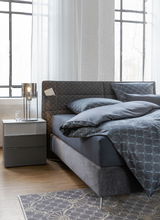 Modernes Bett mit grauem Polsterkopfteil und passender Bettwäsche, seitliche Perspektive mit Nachttisch und Lampe.