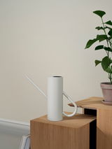 Moderne, minimalistische Blumengießkanne in zylindrischer Form, 2 Liter, in sanftem Weiß, auf einem Holztisch neben einer Pflanze, seitliche Perspektive.