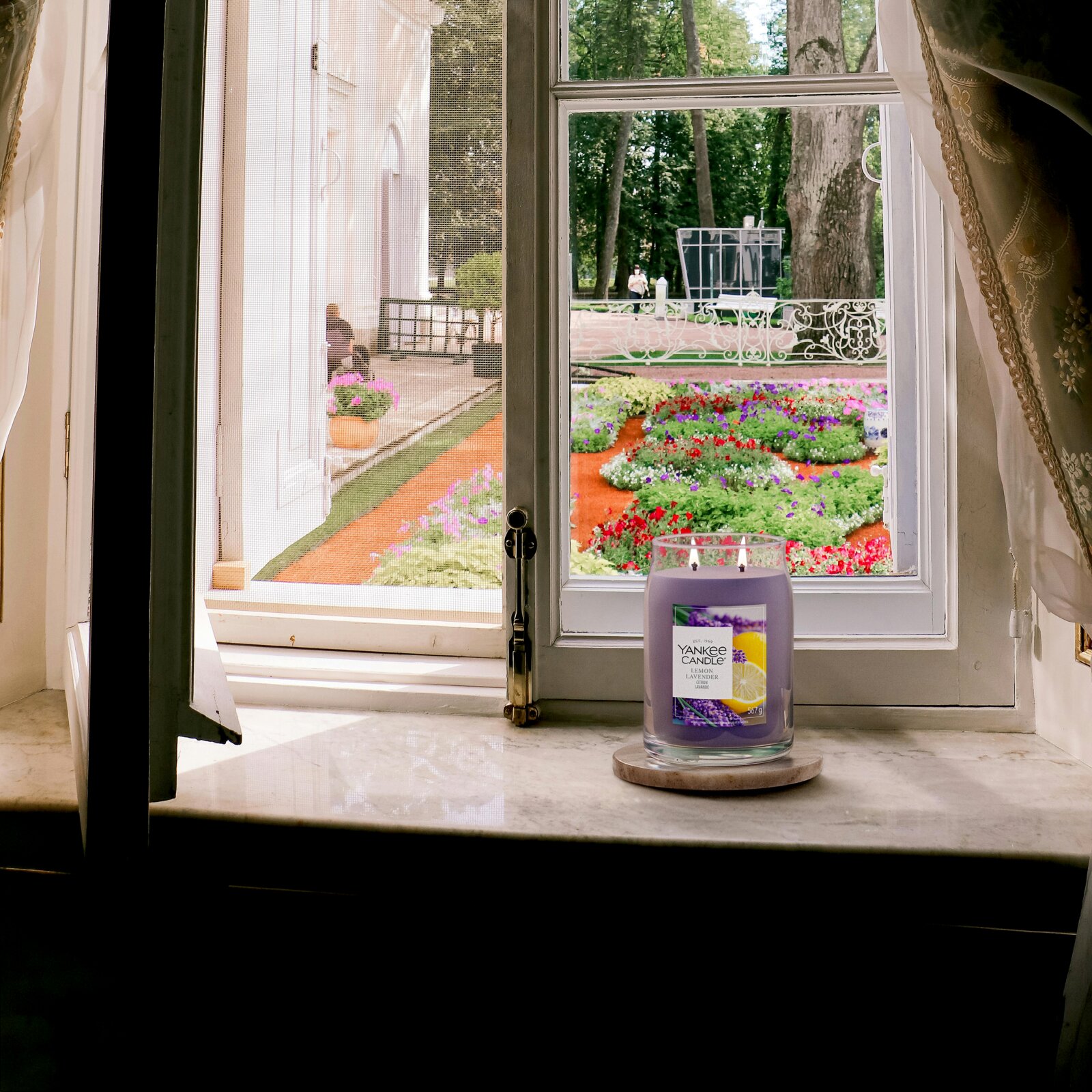 Yankee Candle Lemon Lavender Duftkerze im Glas, frontal auf einer Fensterbank vor einem farbenfrohen Blumengarten präsentiert