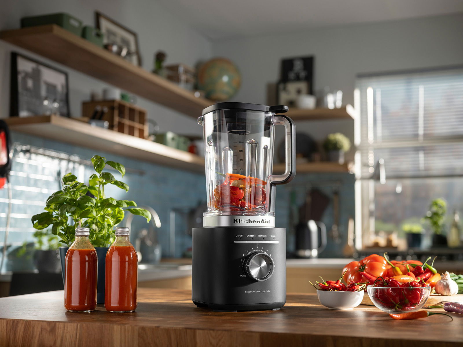 Standmixer in einer modernen Küche, gefüllt mit roten Paprikastücken, umgeben von frischen Zutaten und Kräutern, aus einer seitlichen Perspektive fotografiert.