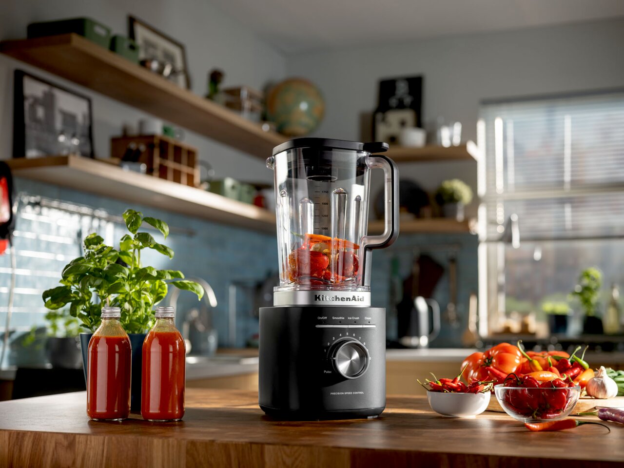 Standmixer in einer modernen Küche, gefüllt mit roten Paprikastücken, umgeben von frischen Zutaten und Kräutern, aus einer seitlichen Perspektive fotografiert.