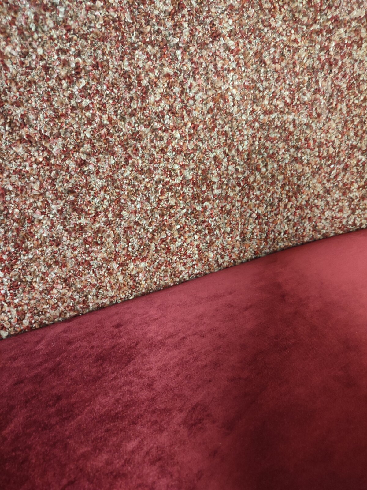 Nahaufnahme in schräger Seitenperspektive: roter, samtiger Polsterstoff vor einer melierten, grob strukturierten Wand- oder Teppichoberfläche in Rot-, Beige- und Brauntönen