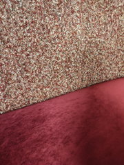 Nahaufnahme in schräger Seitenperspektive: roter, samtiger Polsterstoff vor einer melierten, grob strukturierten Wand- oder Teppichoberfläche in Rot-, Beige- und Brauntönen