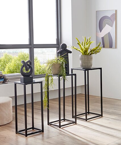 Drei schwarze Metall-Pflanzenständer in unterschiedlichen Höhen im modernen Wohnraum am Fenster, mit Skulpturen und einem Topf mit grüner Pflanze, fotografiert in leichter Schrägansicht von vorn.