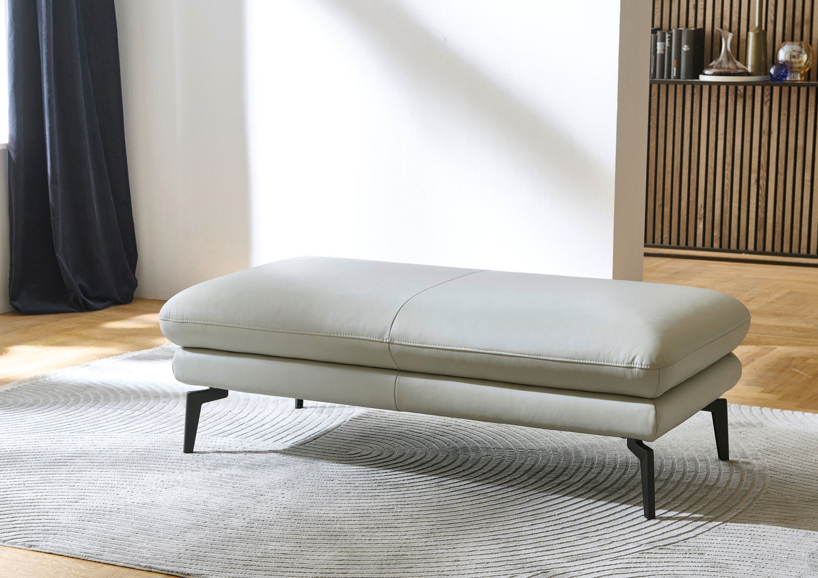 XXL-Hocker in hellgrauem Leder, seitliche Perspektive, auf einem gemusterten Teppich stehend.