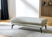 XXL-Hocker in hellgrauem Leder, seitliche Perspektive, auf einem gemusterten Teppich stehend.