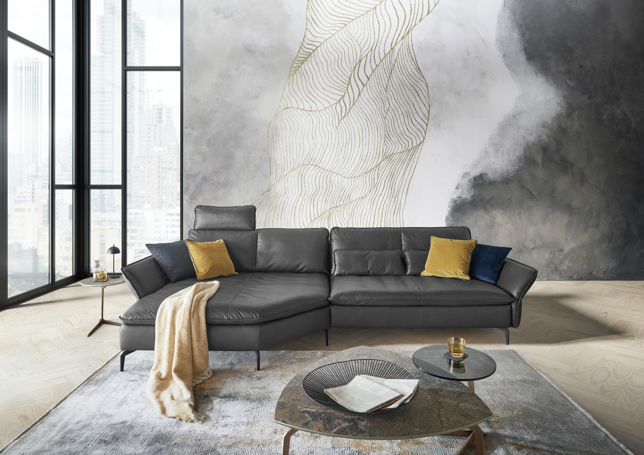 Moderne Longchairkombination in dunklem Leder mit gelben und blauen Kissen, vor einer stilvollen Wand mit abstraktem Muster, aus der Frontalperspektive fotografiert.