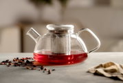 Transparente Glas-Teekanne mit Deckel und Edelstahl-Teesieb, gefüllt mit rotem Tee, auf hellem Tisch mit losen Teeblättern im Vordergrund; seitliche Perspektive auf das Produkt.