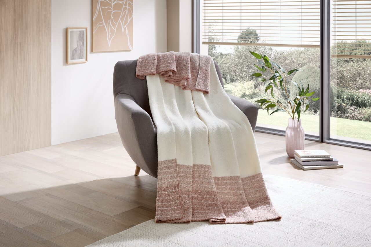 Een gezellige deken in crème en roze op een grijze fauteuil, met uitzicht op een lichte, moderne kamer met grote ramen.