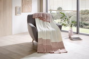 Een gezellige deken in crème en roze op een grijze fauteuil, met uitzicht op een lichte, moderne kamer met grote ramen.