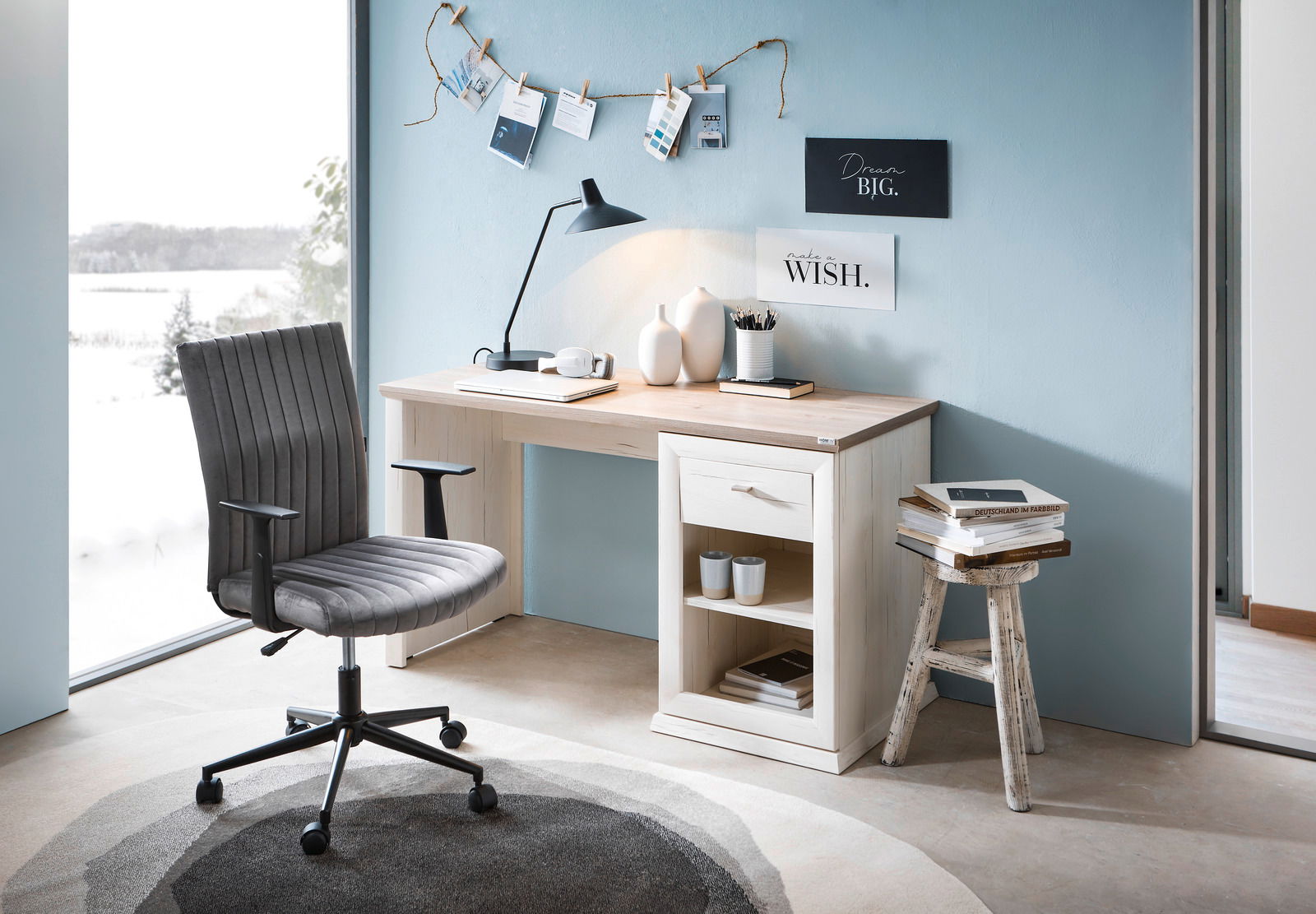 Moderner Schreibtisch in heller Holzoptik mit Schublade und Regal, seitliche Perspektive, dekoriert mit Lampe, Vasen und Büchern, neben einem großen Fenster mit Blick auf eine verschneite Landschaft.