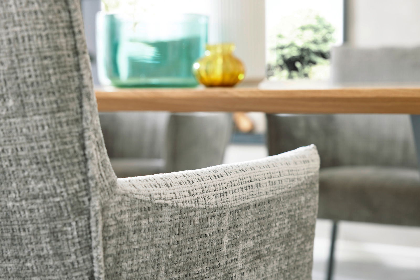 Nahaufnahme eines grauen Armlehnstuhls mit strukturierter Oberfläche, im Hintergrund ein Holztisch mit Dekorationen. Perspektive von der Seite.