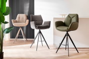 Drei moderne Barstühle in verschiedenen Farben und Materialien, von links nach rechts: beige Stoff, grauer Stoff und grünes Leder, auf einem hellen Holzboden vor einer weißen und dunklen Wand. Perspektive: Frontalansicht.