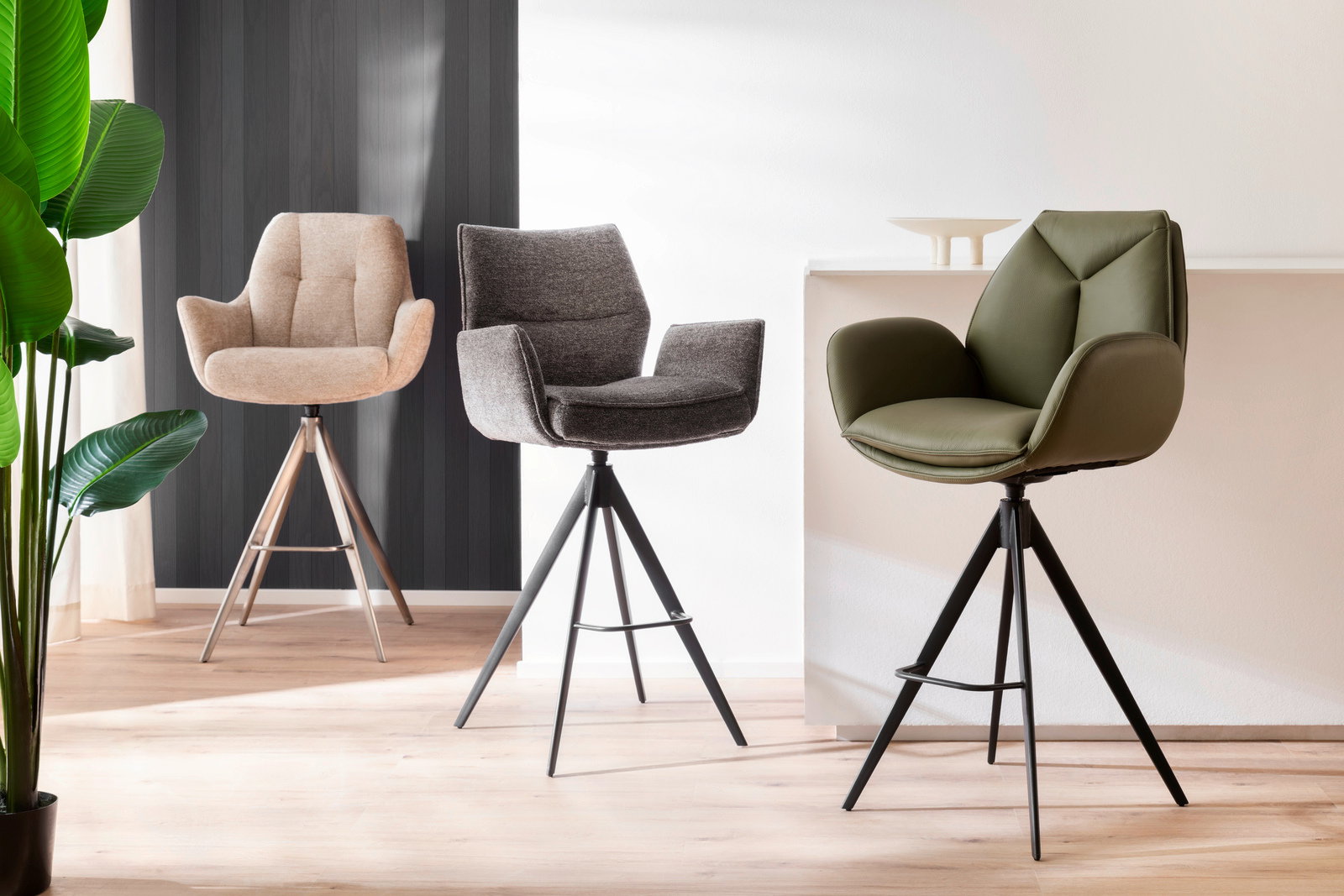 Drei moderne Barstühle in verschiedenen Farben und Materialien, von links nach rechts: beige Stoff, grauer Stoff und grünes Leder, auf einem hellen Holzboden vor einer weißen und dunklen Wand. Perspektive: Frontalansicht.