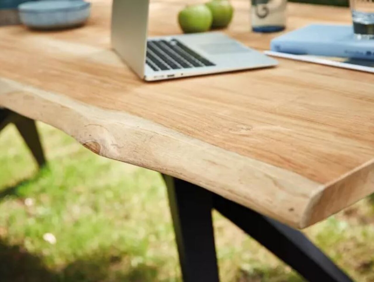 Nahaufnahme eines Esstisches aus Holz mit natürlicher Kante, darauf ein Laptop und grüne Äpfel, im Freien aufgestellt.