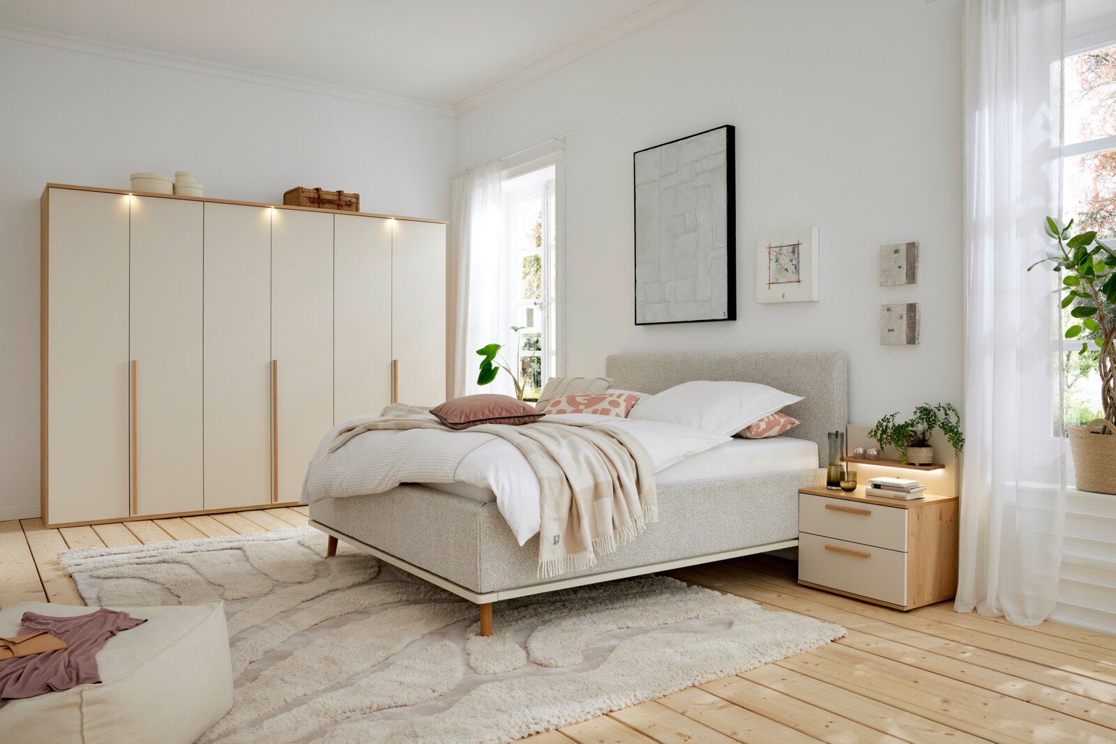 Een moderne slaapkamer met een gestoffeerd bed in een lichte stof, geflankeerd door een nachtkastje en een grote kledingkast op de achtergrond. Het perspectief toont het bed vanaf de zijkant, met een raam dat natuurlijk licht binnenlaat.