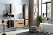 Moderne badkamerset met een aan de muur bevestigde wastafelonderkast in houtlook en antraciet, een witte wastafel, een spiegelkast met ledverlichting en een hoge kast naast het bad bij het raam; perspectief: vooraanzicht van de ruimte, licht van rechts.