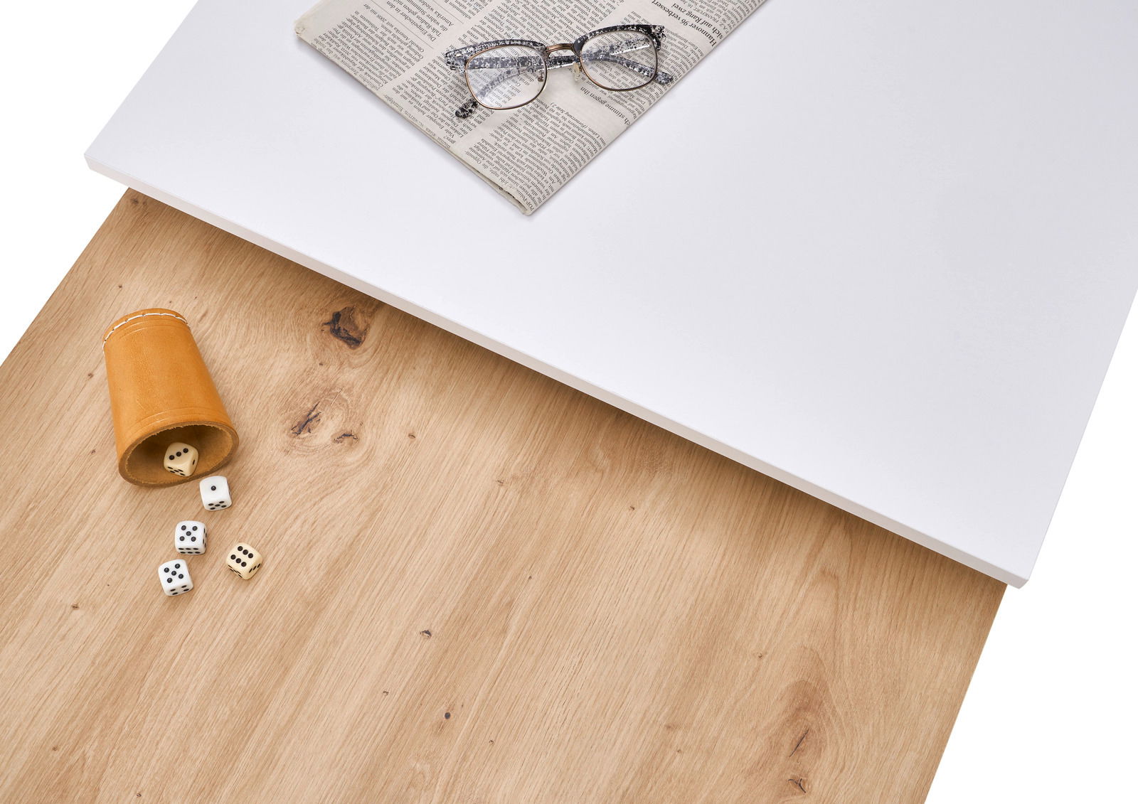 Vogelperspektive auf einen Couchtisch mit einer weißen und einer holzfarbenen Oberfläche, darauf liegen eine Zeitung, eine Brille und ein Würfelbecher mit Würfeln.