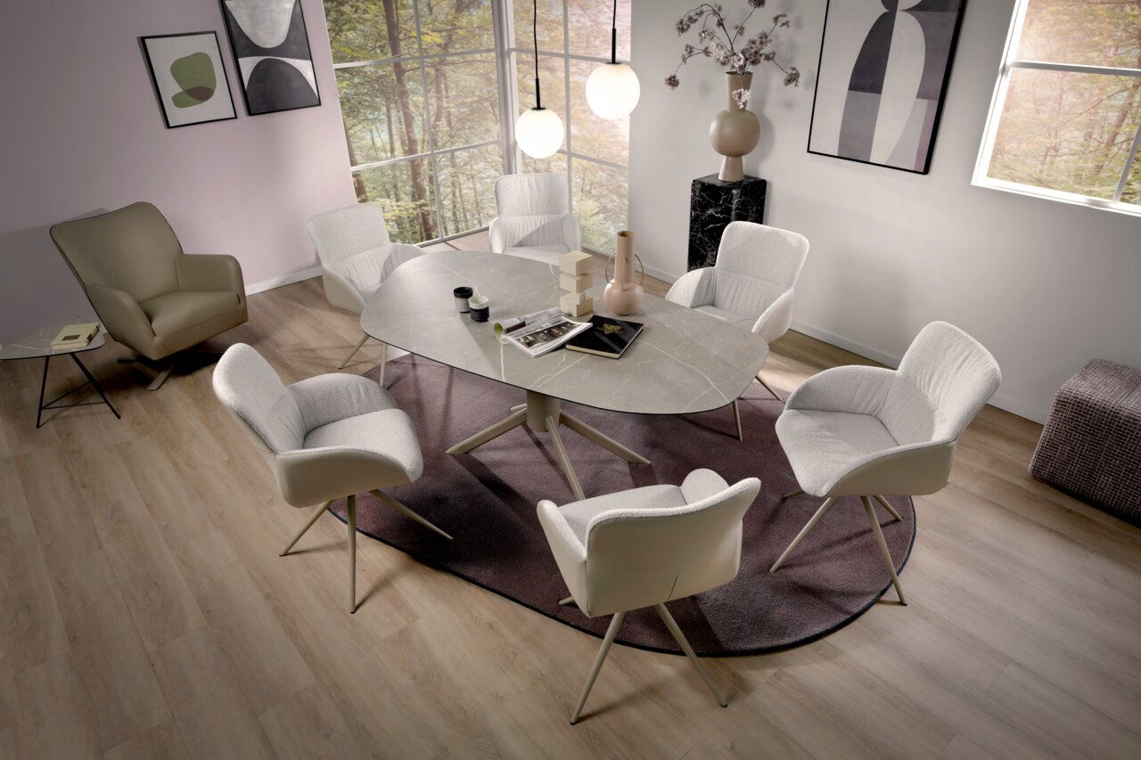 Plantagie Armlehnstuhl RUBY Esszimmer mit modernem Design: Ein ovaler Tisch mit Marmoroptik, umgeben von sechs weißen Armlehnstühlen, auf einem lila Teppich. Im Hintergrund ein großes Fenster mit Blick auf Bäume. Perspektive von oben.