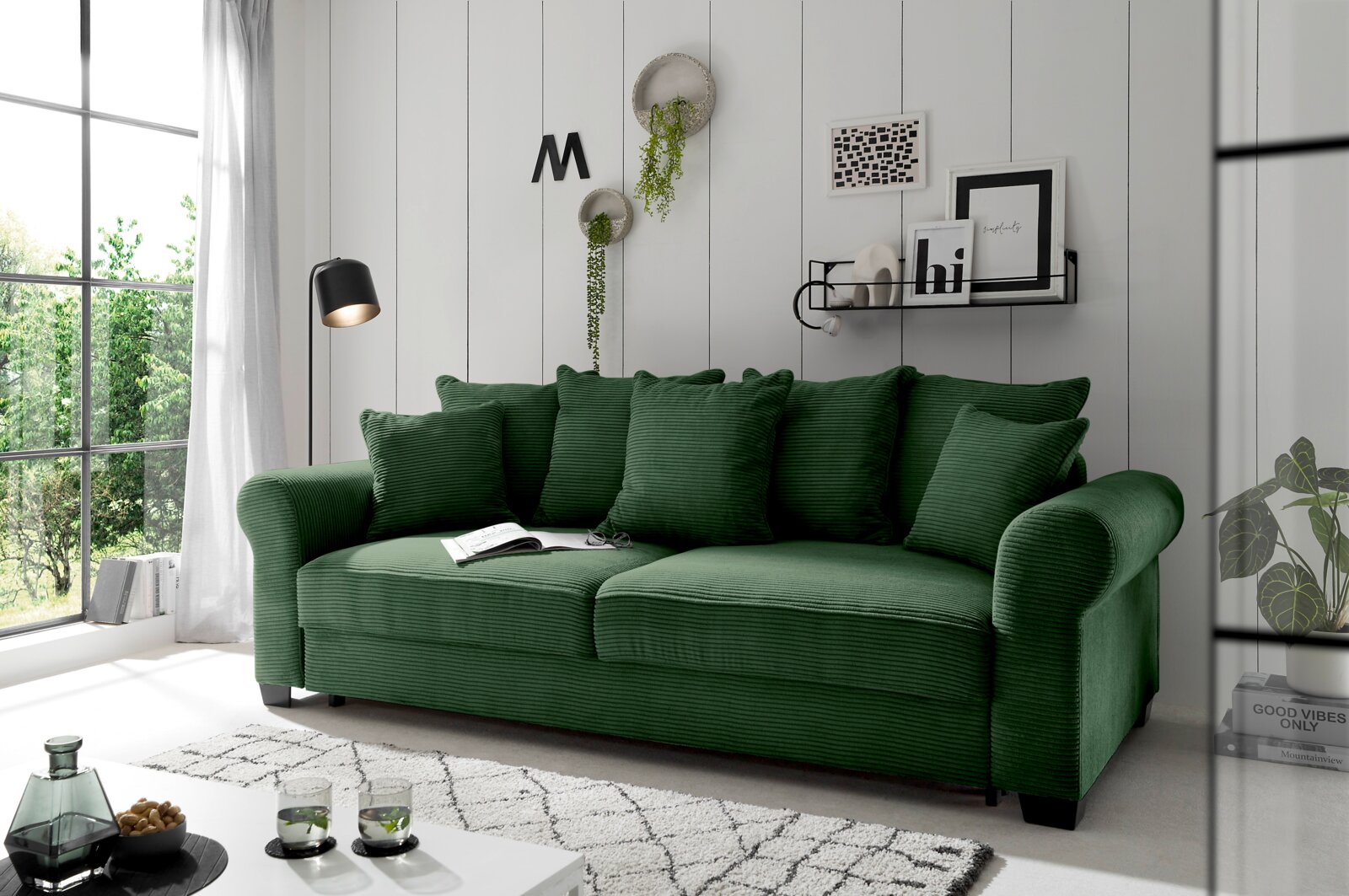 Grünes Funktionssofa mit mehreren Kissen, seitliche Perspektive, in einem modernen Wohnzimmer mit großen Fenstern und stilvoller Dekoration.