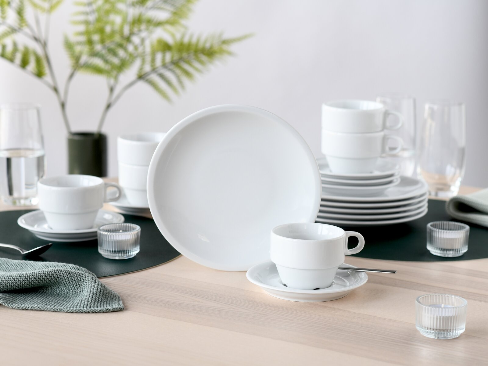 creaTable Kaffeeservice 18-tlg MILANO Weißes Porzellan-Geschirrset mit großem Teller im Vordergrund, Tassen mit Untertassen und gestapelten Tellern auf einem gedeckten Tisch, fotografiert in frontaler Perspektive mit leichtem Blick von vorn.