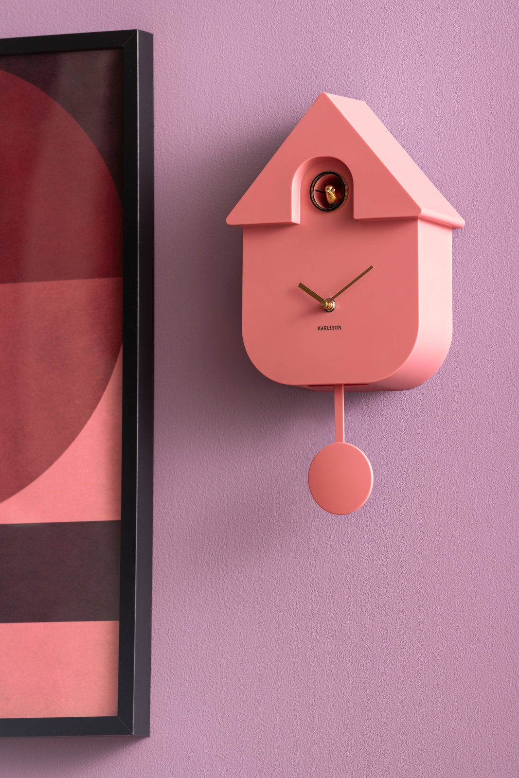 Moderne rosa Wanduhr in Hausform mit Pendel, seitliche Perspektive, an einer lila Wand neben einem gerahmten Bild.