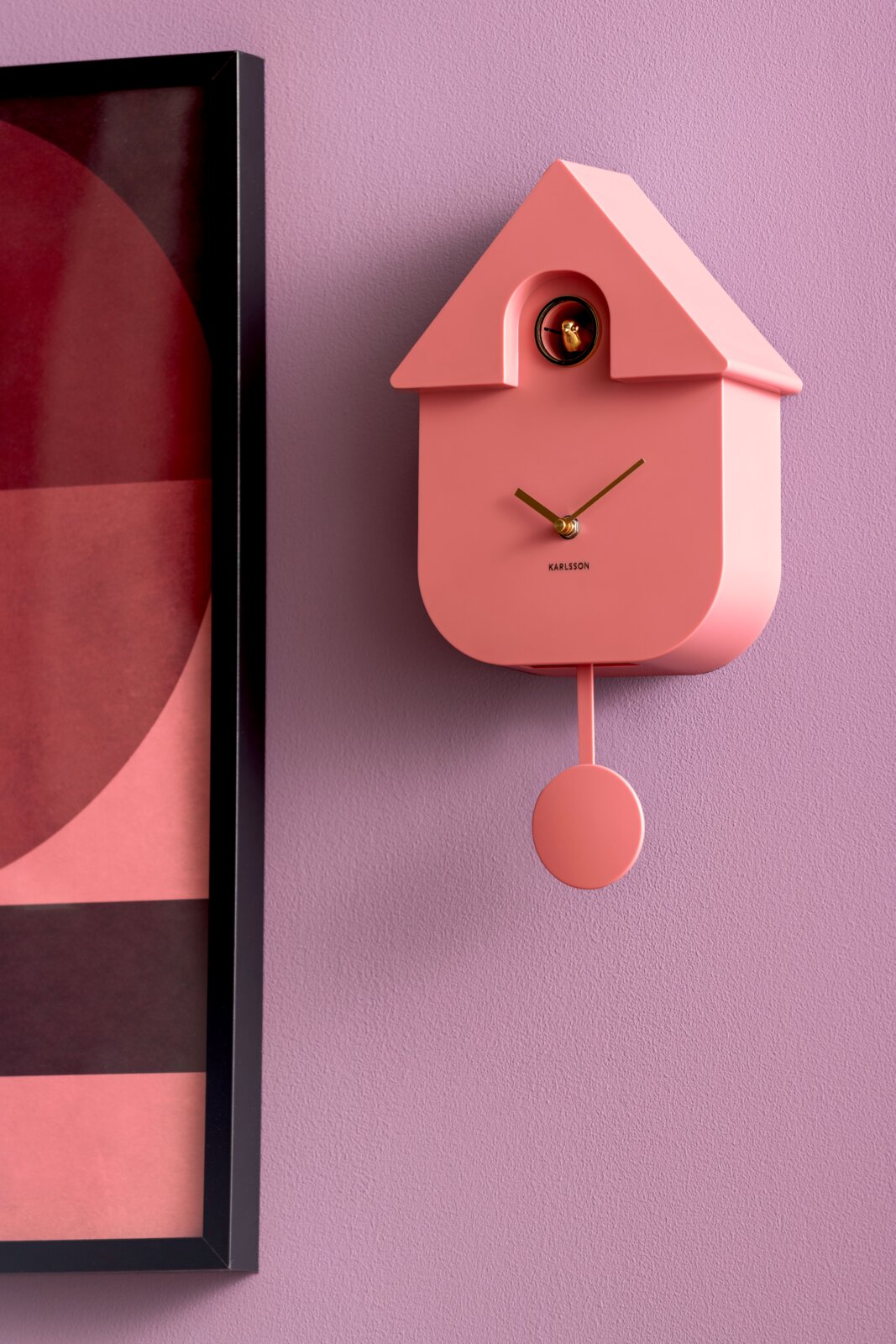 Moderne rosa Wanduhr in Hausform mit Pendel, seitliche Perspektive, an einer lila Wand neben einem gerahmten Bild.