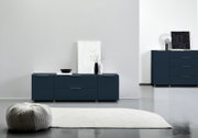 Modernes TV-Lowboard in dunklem Blau mit klaren Linien, frontal fotografiert. Dekoriert mit einer Vase und Kunstobjekten, steht es auf einem glänzenden Boden neben einem Teppich.