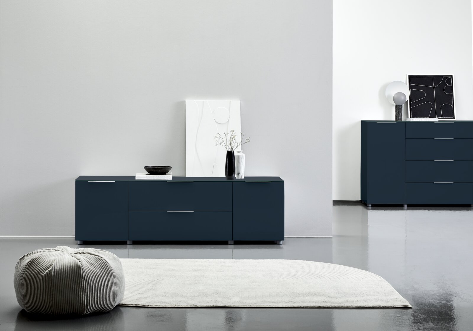 Voleo TV-Lowboard MALIN Modernes TV-Lowboard in dunklem Blau mit klaren Linien, frontal fotografiert. Dekoriert mit einer Vase und Kunstobjekten, steht es auf einem glänzenden Boden neben einem Teppich.