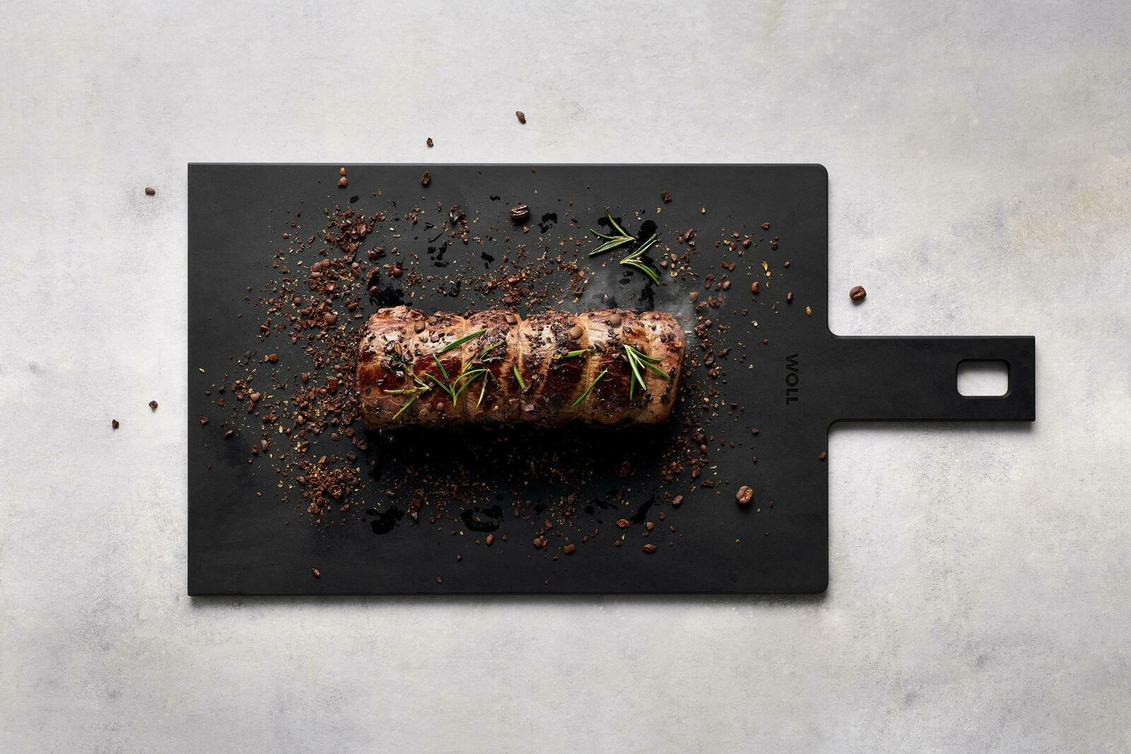 Schwarzes Schneidebrett mit Griff, darauf ein gebratenes Stück Fleisch mit Rosmarin und Gewürzen, aus der Vogelperspektive fotografiert.