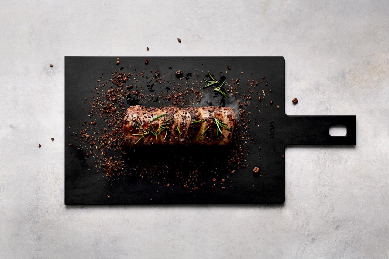 Schwarzes Schneidebrett mit Griff, darauf ein gebratenes Stück Fleisch mit Rosmarin und Gewürzen, aus der Vogelperspektive fotografiert.