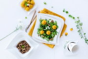 Draufsicht auf einen frischen Blattsalat mit gelben Bällchen (Falafel/Käsebällchen), Walnüssen und weißen Blüten auf einem weißen, eckigen Teller; daneben Schälchen mit weiteren Bällchen und Walnüssen sowie eine Tasse Kaffee auf hellem Hintergrund.