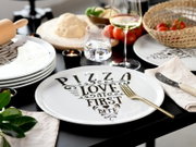 Schwarzer Esstisch mit weißem Pizzateller mit Aufdruck „PIZZA LOVE AT FIRST BITE“, daneben goldene Gabel, Teelichter, Teig und Zutaten; Perspektive: leicht von oben in schräger Frontansicht.