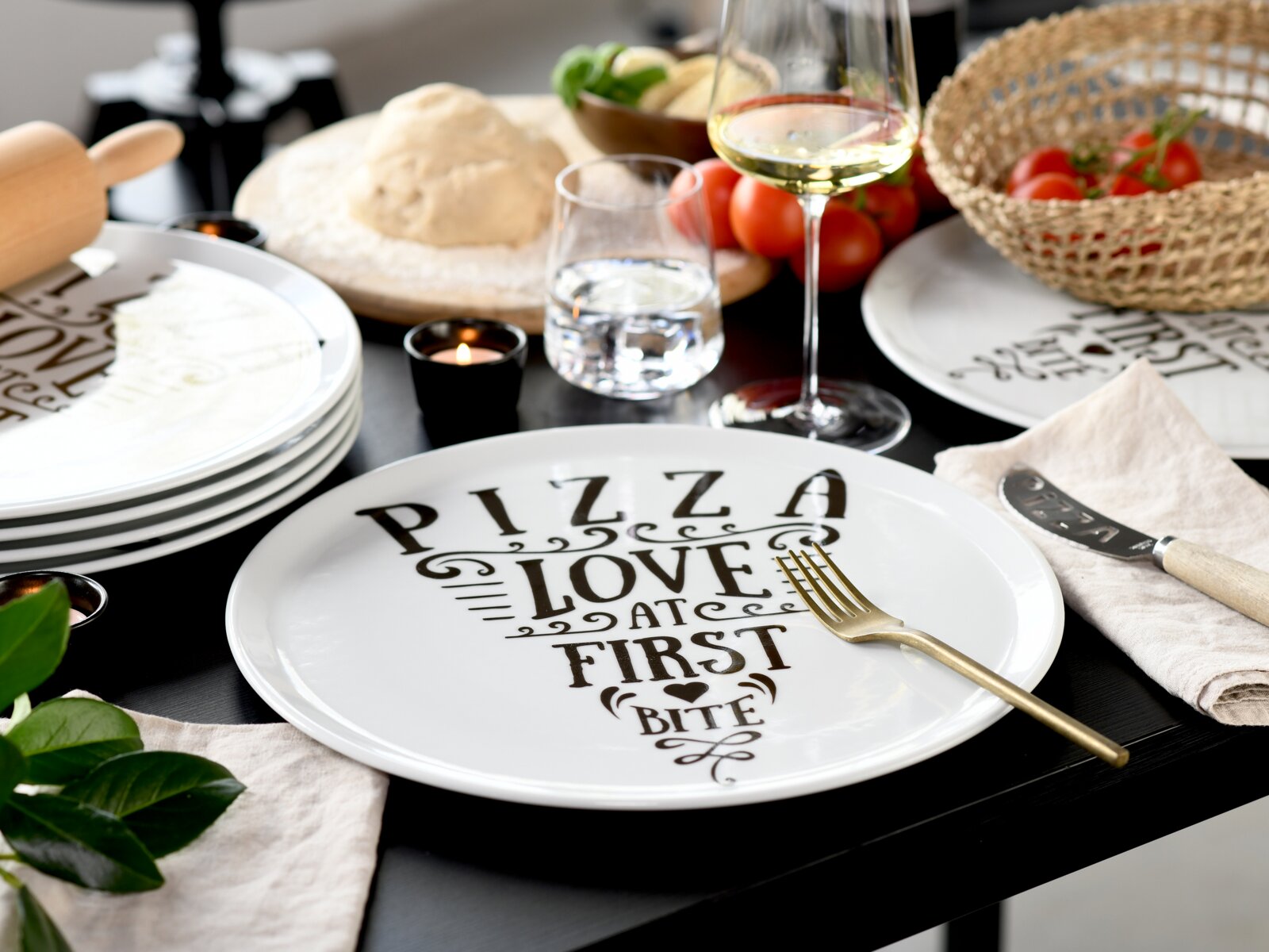 Schwarzer Esstisch mit weißem Pizzateller mit Aufdruck „PIZZA LOVE AT FIRST BITE“, daneben goldene Gabel, Teelichter, Teig und Zutaten; Perspektive: leicht von oben in schräger Frontansicht.