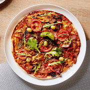 Belegte Pizza mit Salami, Tomatenscheiben, grünen Oliven, Rucola und geschmolzenem Käse auf weißem Teller, von oben (Draufsicht) fotografiert.
