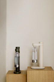 Zwei Wassersprudler in den Farben Olivgrün und Sandbeige, frontal auf Holzwürfeln präsentiert.