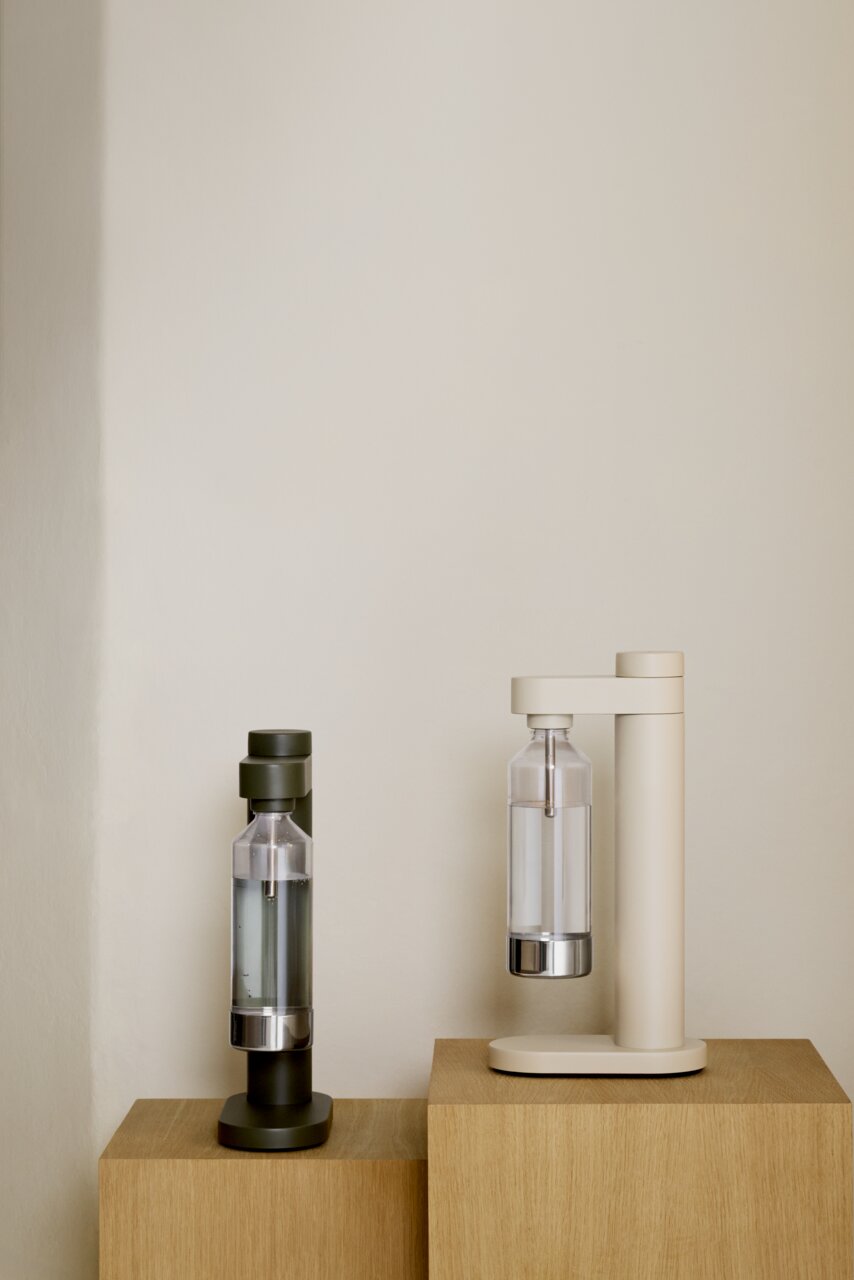 Zwei Wassersprudler in den Farben Olivgrün und Sandbeige, frontal auf Holzwürfeln präsentiert.