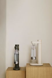 Zwei Wassersprudler in den Farben Olivgrün und Sandbeige, frontal auf Holzwürfeln präsentiert.