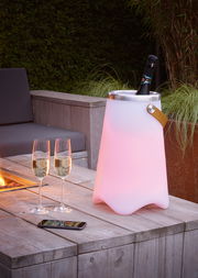 Outdoor-Sektkühler JAMAICA in einem Garten, beleuchtet in sanftem Rosa, mit einer Flasche Sekt und zwei Gläsern auf einem Holztisch, seitliche Perspektive.