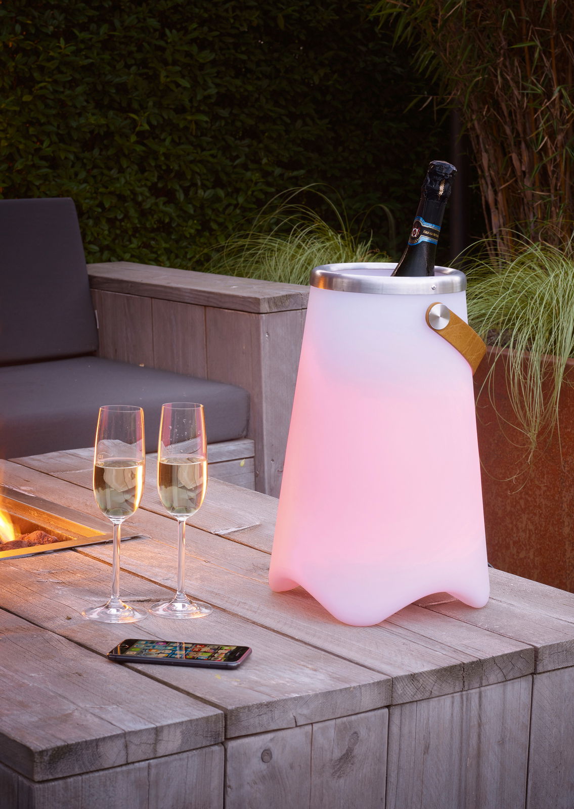 Outdoor-Sektkühler JAMAICA in einem Garten, beleuchtet in sanftem Rosa, mit einer Flasche Sekt und zwei Gläsern auf einem Holztisch, seitliche Perspektive.
