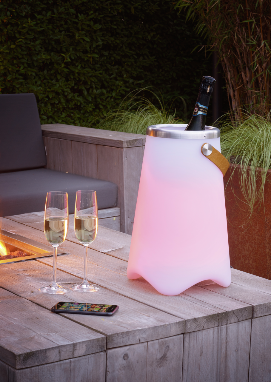 Outdoor-Sektkühler JAMAICA in einem Garten, beleuchtet in sanftem Rosa, mit einer Flasche Sekt und zwei Gläsern auf einem Holztisch, seitliche Perspektive.