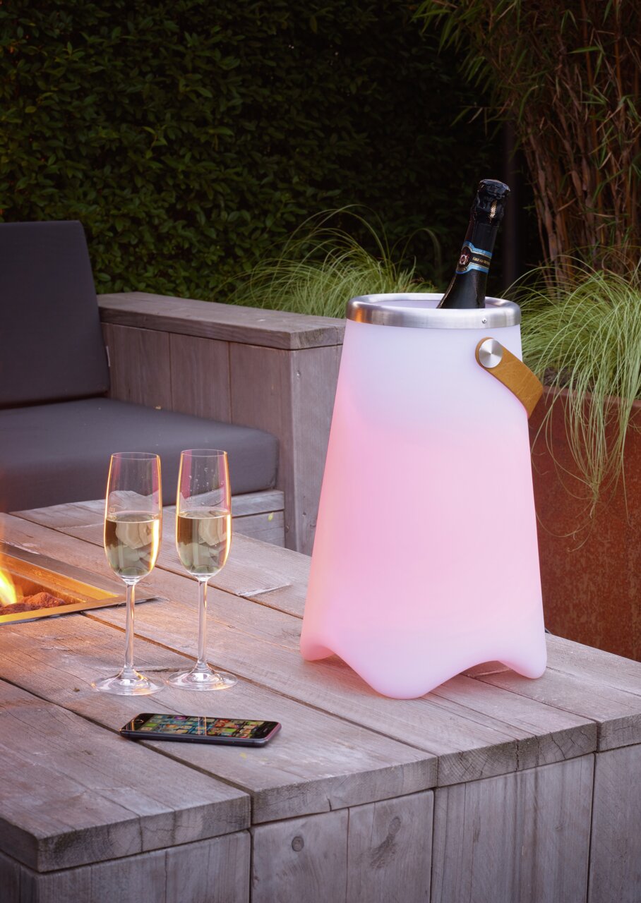 Outdoor-Sektkühler JAMAICA in einem Garten, beleuchtet in sanftem Rosa, mit einer Flasche Sekt und zwei Gläsern auf einem Holztisch, seitliche Perspektive.