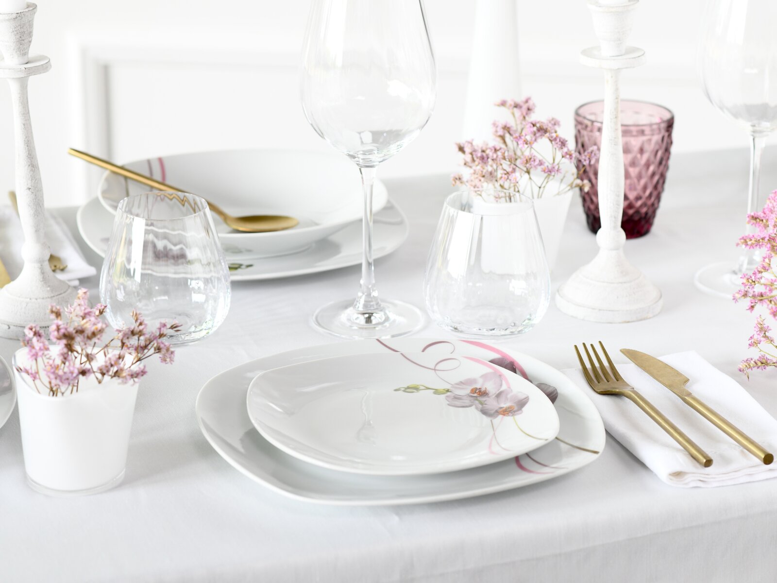 Festlich gedeckter Tisch mit weißem Porzellan-Geschirr mit rosa Blumenmotiv, goldfarbenem Besteck, Wein- und Wassergläsern sowie weißen Kerzenhaltern und rosa Dekoration, aufgenommen in leicht schräger Perspektive von vorne oben