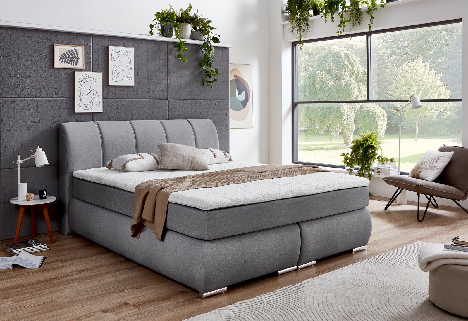 Graues Boxspringbett in einem modernen Schlafzimmer mit großen Fenstern und Pflanzen, seitliche Perspektive.
