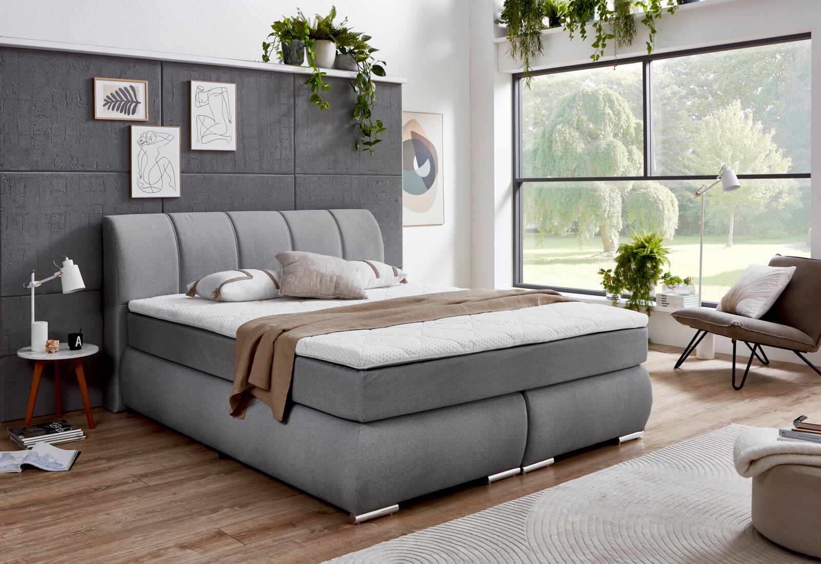 Graues Boxspringbett in einem modernen Schlafzimmer mit großen Fenstern und Pflanzen, seitliche Perspektive.