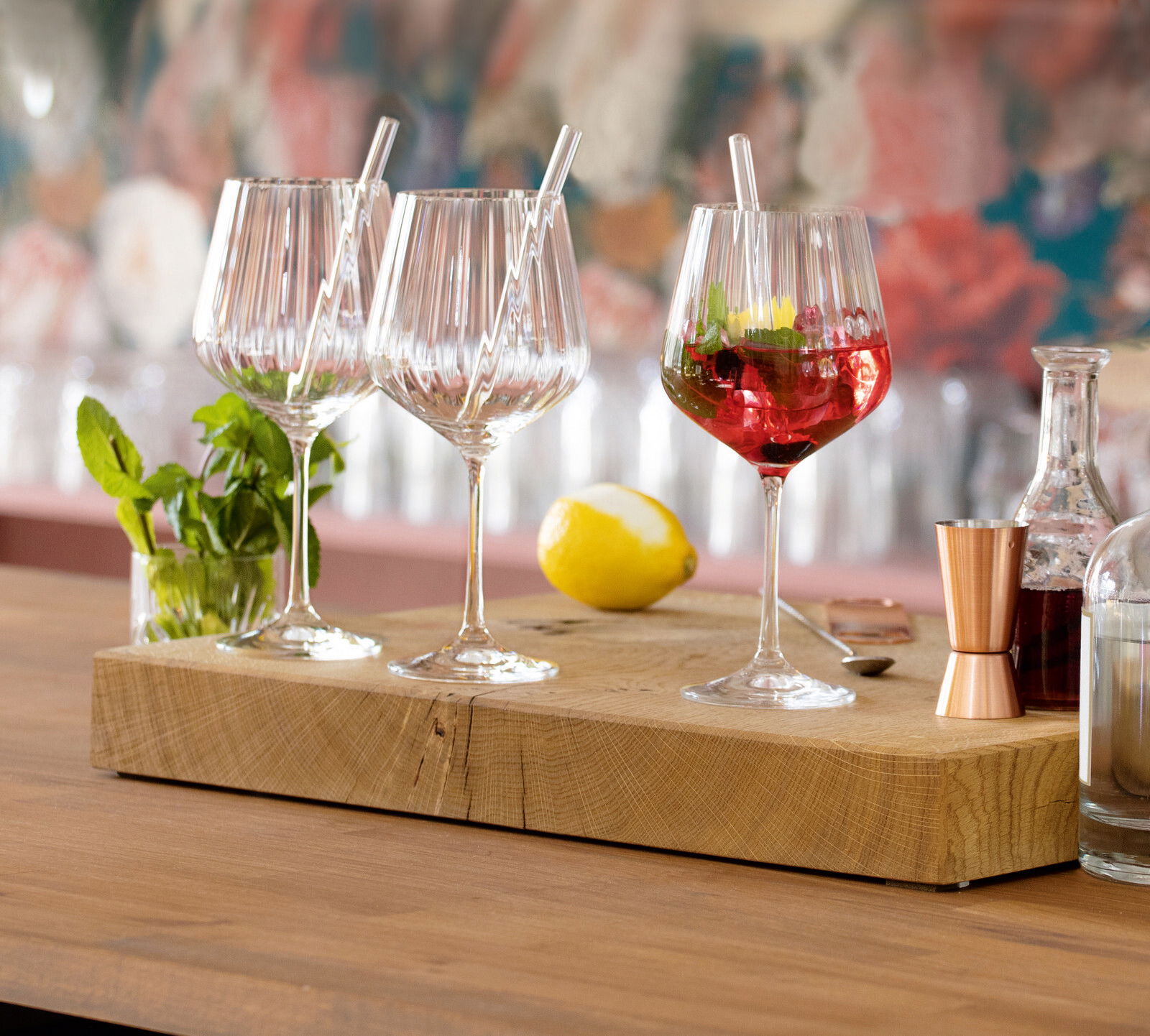 Drei elegante Gin Tonic-Gläser mit Strohhalmen auf einem Holzbrett, umgeben von Minze, Zitrone und Barzubehör, aus seitlicher Perspektive fotografiert.