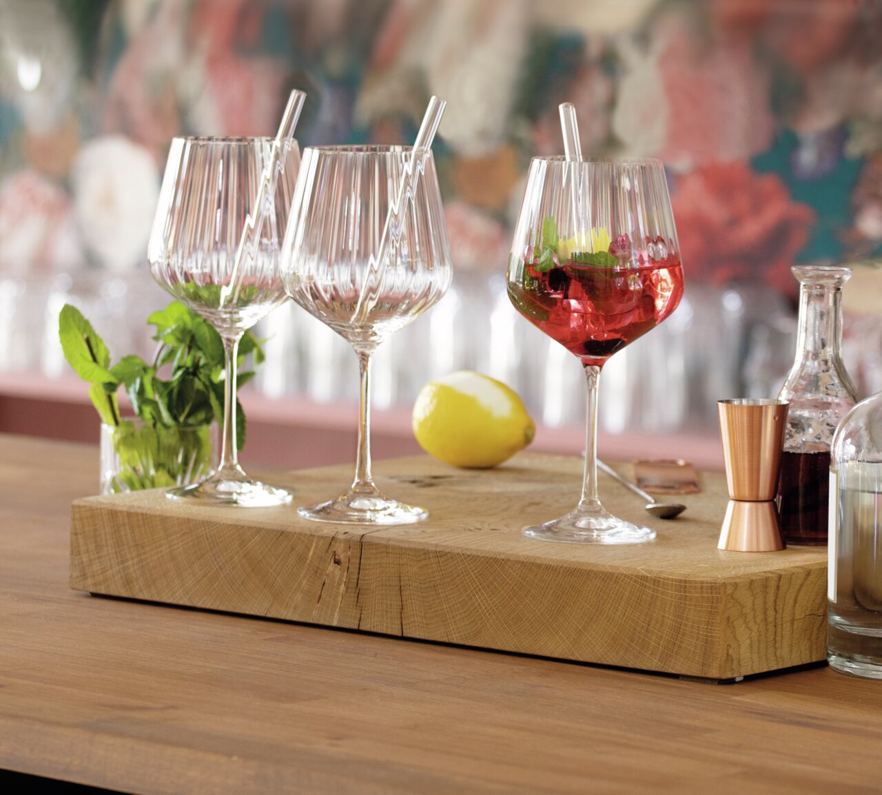 Drei elegante Gin Tonic-Gläser mit Strohhalmen auf einem Holzbrett, umgeben von Minze, Zitrone und Barzubehör, aus seitlicher Perspektive fotografiert.