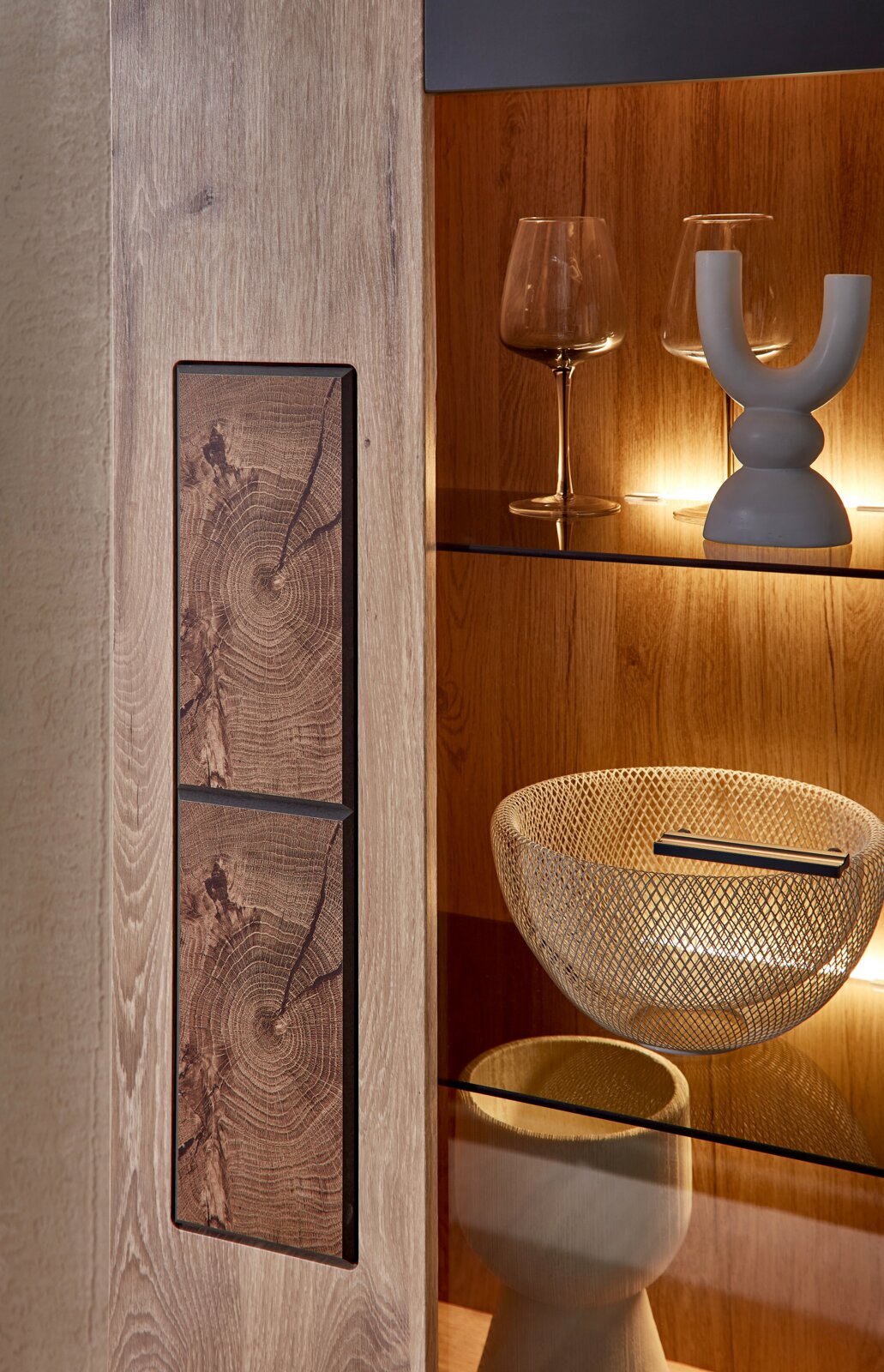 Stylife Vitrine (links) SINA Detailaufnahme in leicht seitlicher Perspektive: Holzschrank/Vitrine mit markanten Holzmaserungs-Fronten und Glasböden, warm beleuchtet, mit Weingläsern und dekorativer Schale.