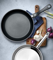 Zwei Pfannen-Set aus der Vogelperspektive, eine mit Antihaftbeschichtung und eine aus Edelstahl, auf einem Schneidebrett mit Zwiebeln und Knoblauch.